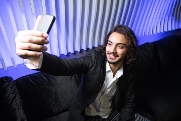 Heureux jeune homme élégant avec smartphone faisant selfie alors qu'il était assis sur un canapé en velours noir en boîte de nuit