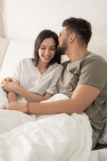 Heureux jeune homme embrassant sa femme souriante au lit