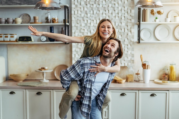 Heureux jeune homme européen roule sa femme sur le dos et s'amuse ensemble dans une cuisine moderne à l'intérieur