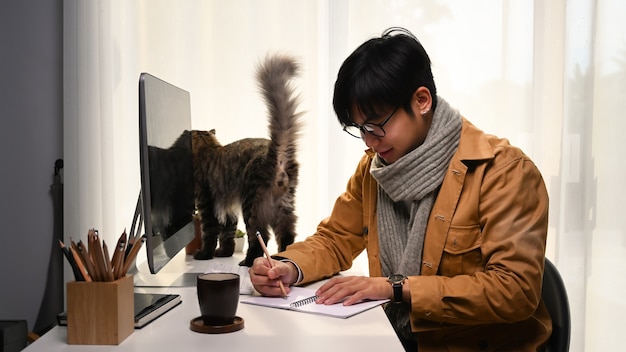 Heureux jeune homme faisant une note sur un ordinateur portable alors qu'il était assis dans une maison confortable avec son chat.