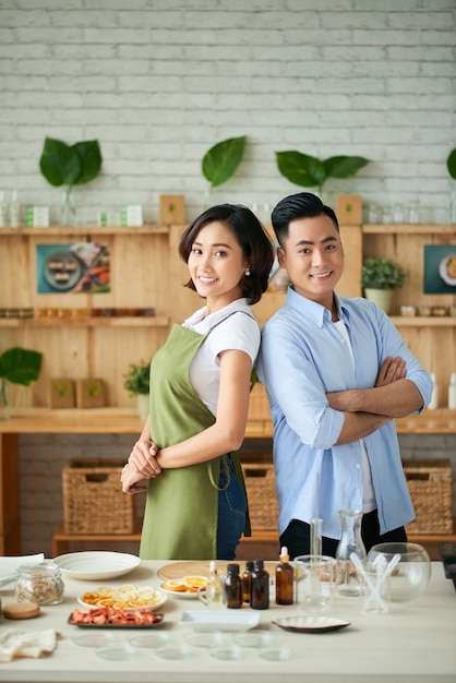 Heureux jeune homme et femme debout dos à dos à table avec des ingrédients pour faire du savon bio et des barres de nettoyage
