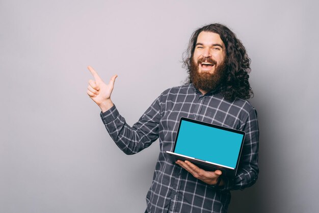 Heureux jeune homme hipster barbu en chemise tenant le doigt pointé de l'ordinateur sur fond gris.