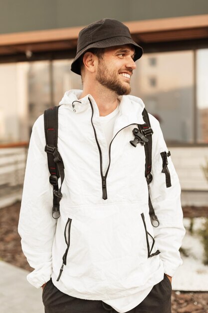 Heureux jeune homme hipster élégant en chapeau de mode avec veste blanche à la mode et sac à dos voyage dans la ville