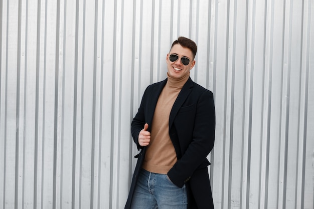 Heureux jeune homme hipster en manteau d'automne élégant en pull tricoté dans des lunettes de soleil à la mode en jeans bleu est debout et montrant les pouces vers le haut près du mur métallique