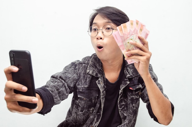 Heureux jeune homme indonésien séduisant choqué joyeusement tout en tenant du papier-monnaie et un téléphone en roupie.