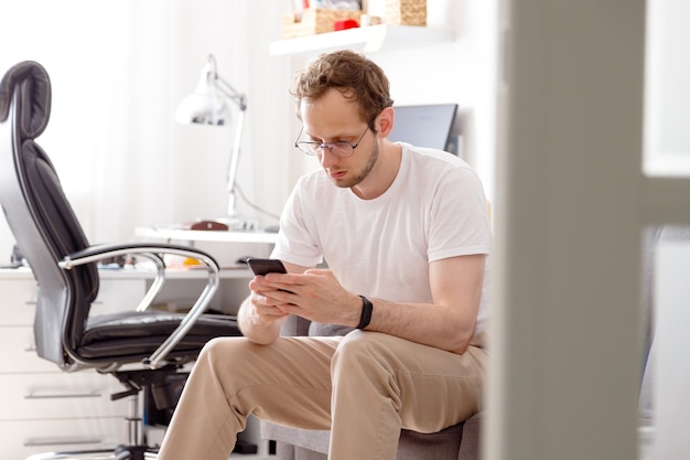 Heureux jeune homme à lunettes assis sur un canapé envoyant du texte sur téléphone mobile