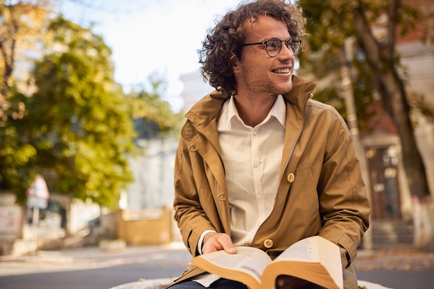 Heureux jeune homme avec des lunettes lisant et posant avec un livre à l'extérieur Étudiant de sexe masculin portant des livres sur le campus dans la rue d'automne