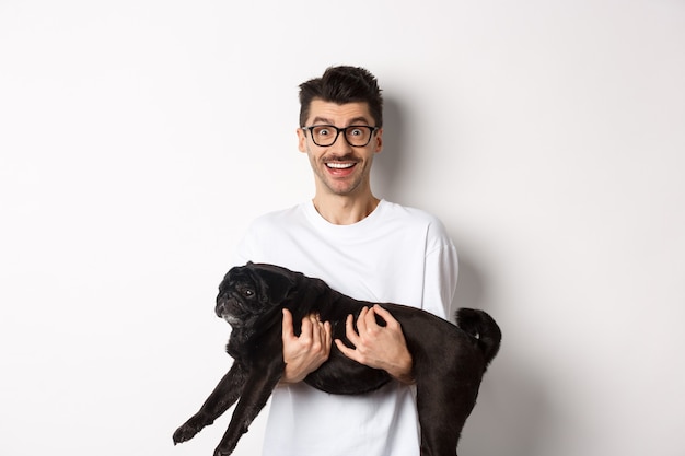 Heureux jeune homme à lunettes tenant un animal de compagnie carlin noir et regardant la caméra amusé, souriant gai, debout sur fond blanc.