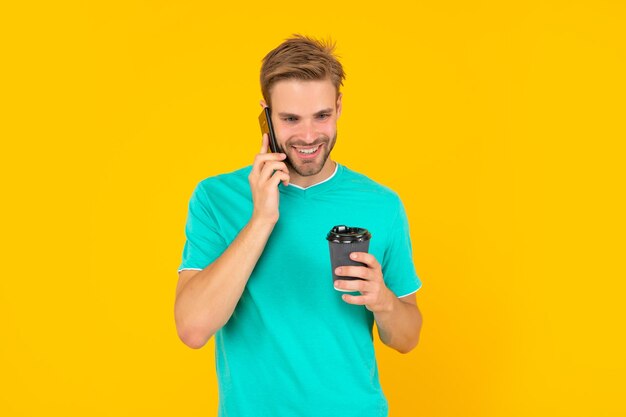 Heureux jeune homme mal rasé parler au téléphone avec une tasse de café sur fond jaune conversation