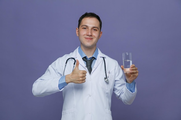 Heureux jeune homme médecin portant une robe médicale et un stéthoscope autour du cou montrant un verre d'eau regardant la caméra montrant le pouce vers le haut isolé sur fond violet