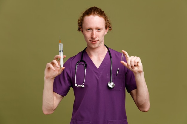 Heureux jeune homme médecin portant l'uniforme avec stéthoscope tenant la seringue avec ampoule isolé sur fond vert