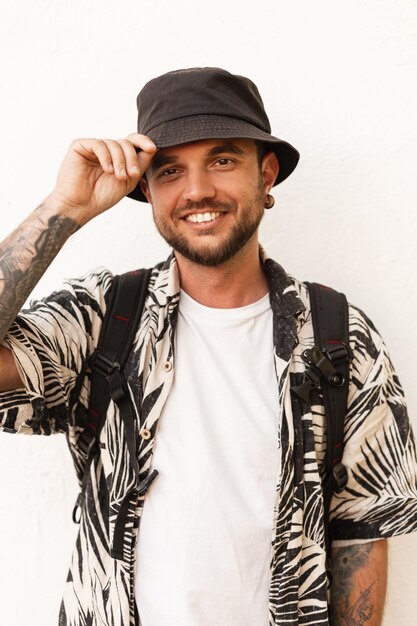 Heureux jeune homme modèle avec sourire dans des vêtements de mode avec chapeau et chemise se tient près du mur blanc