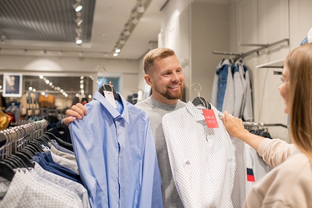 Heureux jeune homme montrant deux chemises à sa petite amie tout en choisissant un nouveau lors de la vente saisonnière dans le département des vêtements décontractés