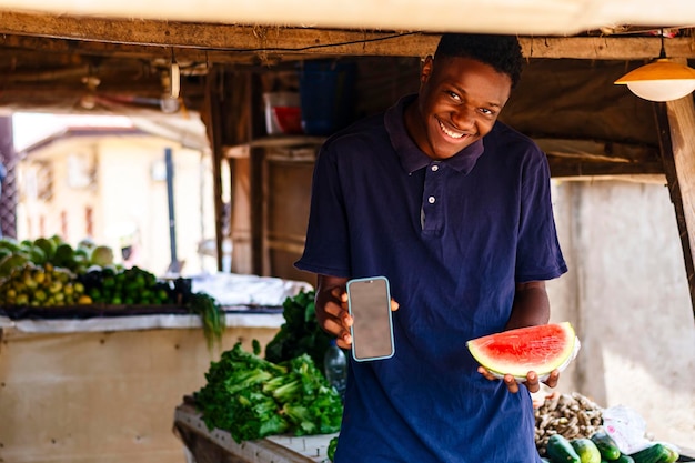 Photo heureux jeune homme noir vendant des stands de fruits avec un smartphone et une pastèque