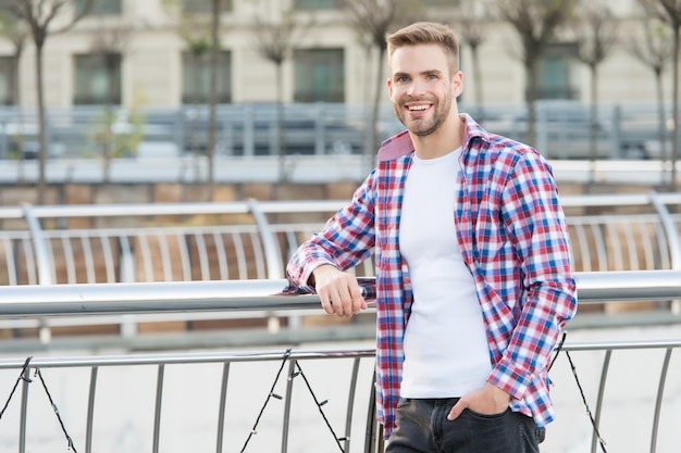 Heureux jeune homme non rasé souriant aux cheveux coiffés en chemise décontractée à carreaux en plein air, mode et coiffeur masculin.