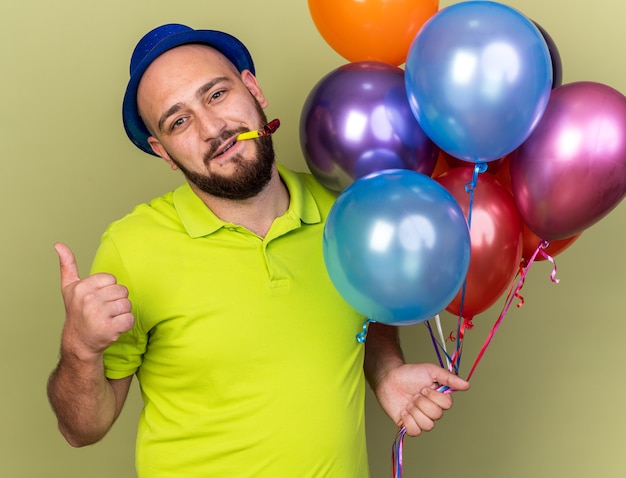 Heureux jeune homme portant un chapeau de fête tenant des ballons soufflant un sifflet de fête montrant le pouce vers le haut isolé sur un mur vert olive