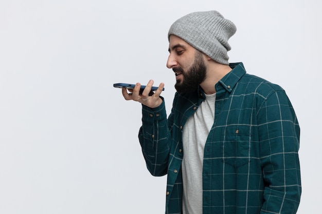 Heureux jeune homme portant un chapeau d'hiver debout dans la vue de profil regardant vers le bas parler au téléphone à l'aide de son microphone isolé sur fond blanc avec espace de copie