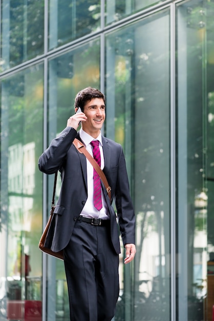 Heureux jeune homme portant un costume tout en parlant au téléphone mobile et en marchant le long d'un bâtiment d'entreprise moderne