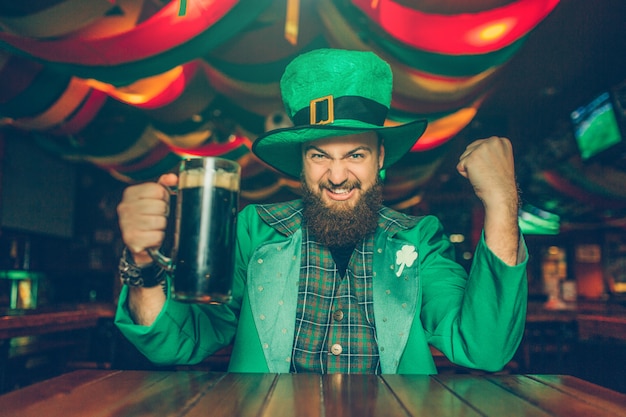 Heureux jeune homme porte le costume de Saint-Patrick. Il est assis à table dans un bar et tient une chope de bière brune. Guy a l'air heureux et excité.
