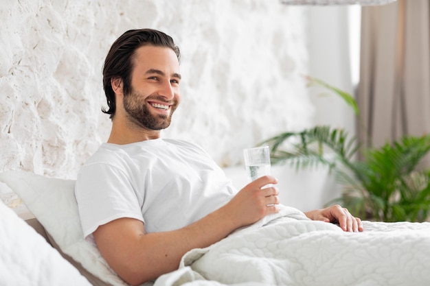 Heureux jeune homme prenant un verre d'eau au lit