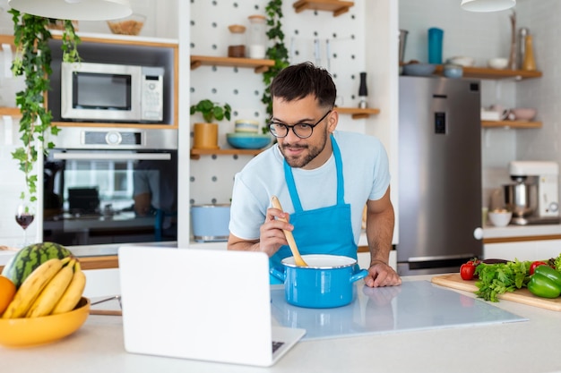 Heureux jeune homme préparant un dîner sain à la maison Il suit un didacticiel vidéo sur l'ordinateur portable