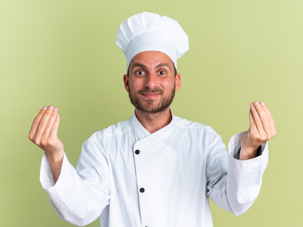 Heureux jeune homme de race blanche cuisinier en uniforme de chef et casquette regardant la caméra faire semblant de tenir quelque chose dans les mains isolé sur un mur vert olive