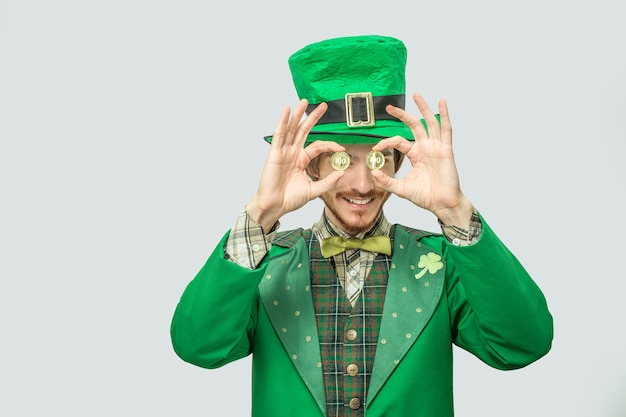 Heureux jeune homme riche en costume vert tenir deux pièces d'or devant les yeux. Il sourit et a l'air heureux. Guy porte le costume vert de Saint Patrick. Isolé sur gris.