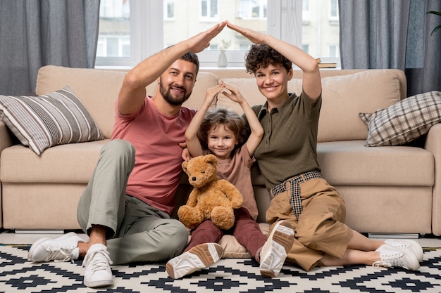 Heureux jeune homme, sa femme et leur mignon petit fils d'âge élémentaire gardant les bras au-dessus de la tête et faisant le toit assis sur le sol