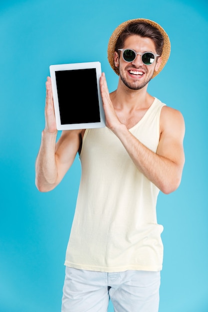 Heureux jeune homme séduisant au chapeau et lunettes de soleil tenant une tablette à écran vierge sur un mur bleu