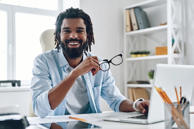 Heureux jeune homme souriant et regardant la caméra tout en travaillant à l'intérieur