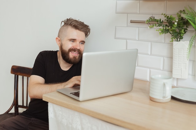 Heureux jeune homme souriant regardant et travaillant sur un ordinateur portable à la maison. Technologies, télétravail et réseaux sociaux