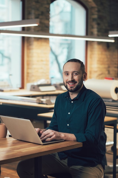 Heureux jeune homme à la table souriant et vous regardant assis avec ses mains sur le clavier de l'ordinateur portable