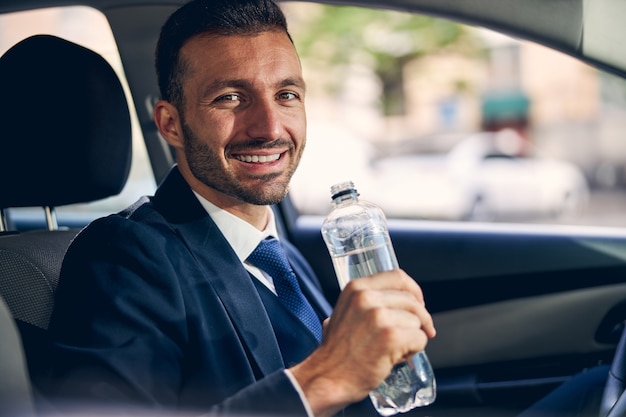 Heureux jeune homme tenant une bouteille d'eau et regardant directement la caméra alors qu'il était assis sur sa voiture