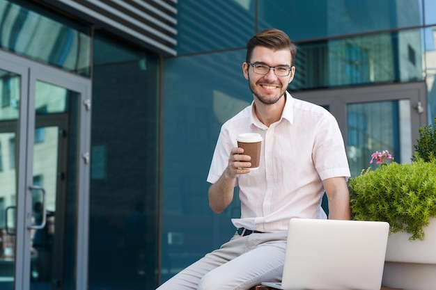 Heureux jeune homme travaillant sur un ordinateur portable et buvant du café assis sur un banc près d'un immeuble de bureaux moderne, vue de face, espace de copie. Concept d'entreprise, de technologie et de réussite