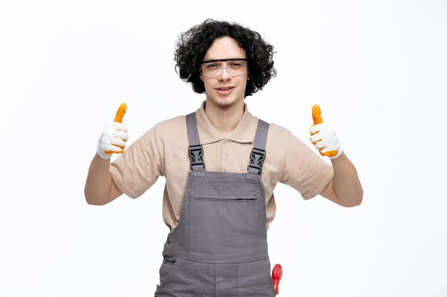 Heureux jeune homme travailleur de la construction portant des lunettes de sécurité uniformes et des gants de sécurité regardant la caméra montrant les pouces vers le haut isolé sur fond blanc