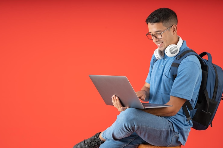 Photo heureux jeune homme d'université avec des lunettes et des écouteurs blancs sur son cou assis à l'aide de son ordinateur portable