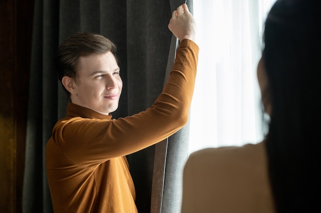 Heureux jeune homme en vêtements décontractés intelligents regardant sa femme debout devant lui tout en dessinant des rideaux sombres dans une chambre d'hôtel le soir