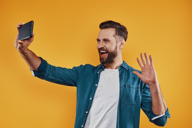 Heureux jeune homme en vêtements décontractés prenant selfie à l'aide d'un téléphone intelligent et souriant en se tenant debout
