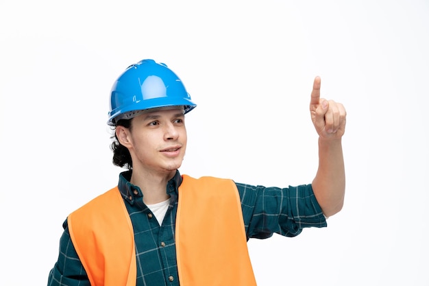 Heureux jeune ingénieur masculin portant un casque de sécurité et un gilet de sécurité regardant le côté pointant le doigt vers le haut isolé sur fond blanc