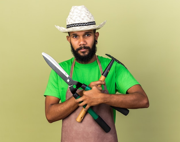 Heureux jeune jardinier afro-américain portant un chapeau de jardinage tenant et traversant des tondeuses avec un râteau isolé sur un mur vert olive