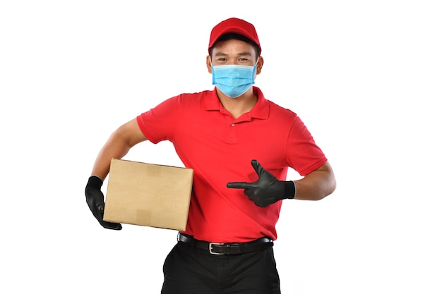Heureux jeune livreur asiatique en uniforme rouge, masque médical, gants de protection transportent une boîte en carton dans les mains isolés sur blanc. Le livreur donne l'envoi de colis. Livraison sûre