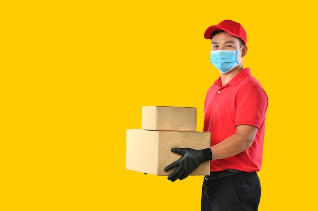 Heureux jeune livreur asiatique en uniforme rouge, masque médical, gants de protection transportent une boîte en carton dans les mains sur le mur jaune. Le livreur donne l'envoi de colis. Pendant l'épidémie de COVID-19