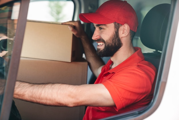 Heureux jeune livreur en uniforme rouge souriant et conduisant une camionnette avec des boîtes à colis