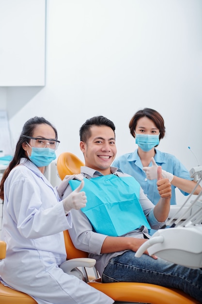 Heureux jeune patient asiatique, son dentiste et son assistant montrant le pouce levé et souriant à la caméra après un traitement dentaire