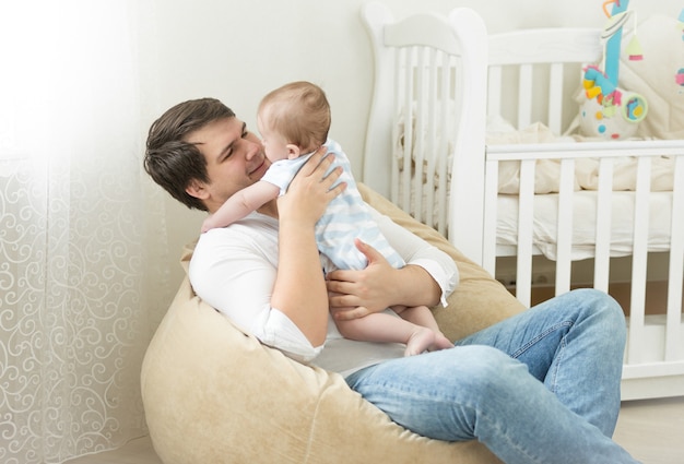 Heureux jeune père assis dans un sac et jouant avec son fils de 6 mois