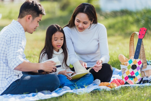 Heureux jeune père de famille asiatique, mère et enfant petite fille s'amusant et profitant de l'extérieur sur une couverture de pique-nique lisant un livre dans le parc à l'heure ensoleillée, concept de printemps de loisirs d'été