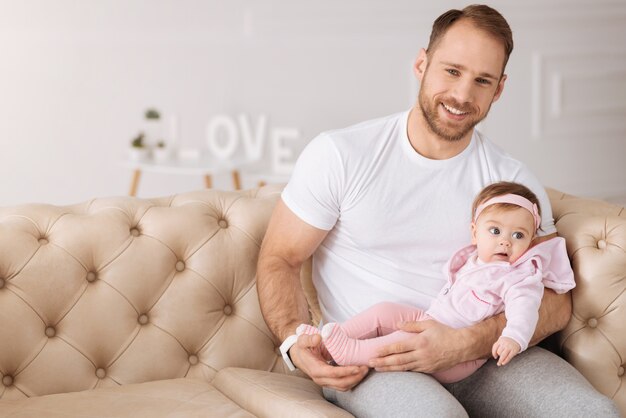 Heureux jeune père. Homme barbu souriant athlétique assis sur le canapé à la maison et tenant son bébé tout en exprimant la positivité