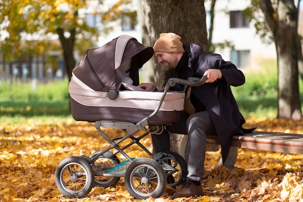 Photo heureux jeune père avec landau assis sur un banc dans la nature au parc. temps de l'automne