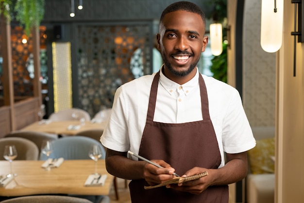 Heureux jeune serveur avec un sourire à pleines dents tenant un stylo et un petit bloc-notes tout en se tenant devant la caméra au restaurant et en prenant les commandes