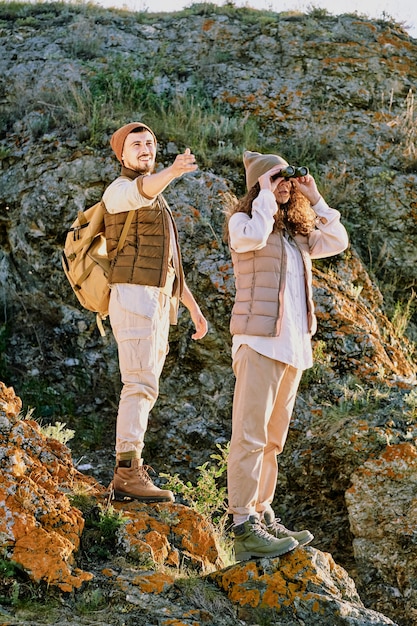 Heureux jeune touriste avec sac à dos pointant vers l'avant en se tenant debout sur un rocher derrière sa femme regardant à travers des jumelles pendant le voyage